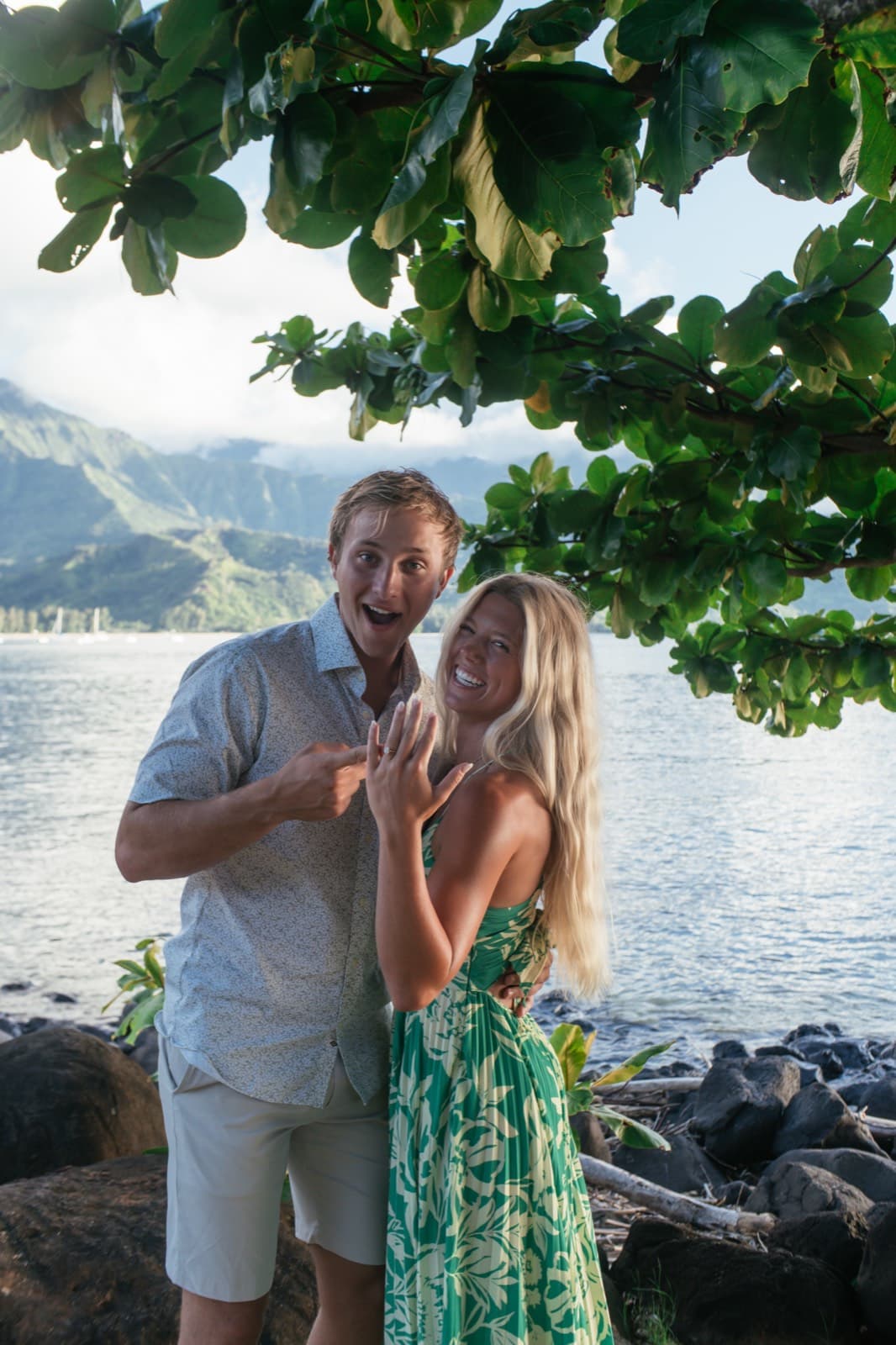 Will points at Emma's engagement ring as they both shout with joy, a bay and mountains behind them