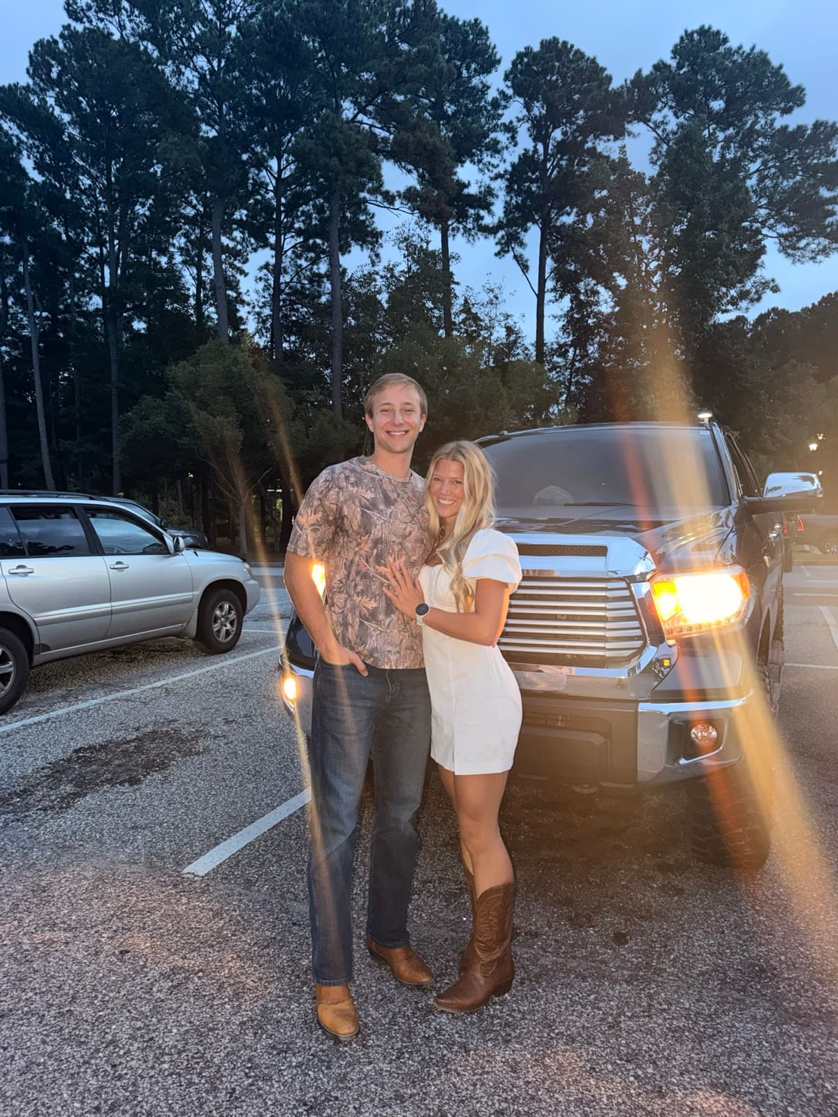 Will and Emma in front of his pickup at dusk under tall pines, headlights flaring